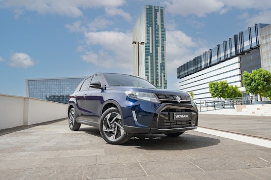 Ein goldgelber Suzuki fährt auf einem Waldweg, umgeben von hohen Bäumen und Sonnenlicht, das durch das Blätterdach fällt.