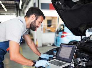 Automechaniker beugt sich in der Werkstatt über den Motorraum eines Fahrzeugs und liest Diagnosedaten von einem angeschlossenen Laptop ab.